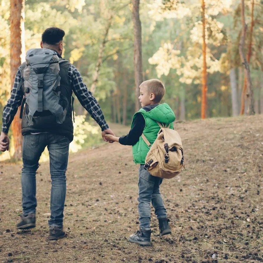 A father and his young son holding hands on a walk through a pine forest